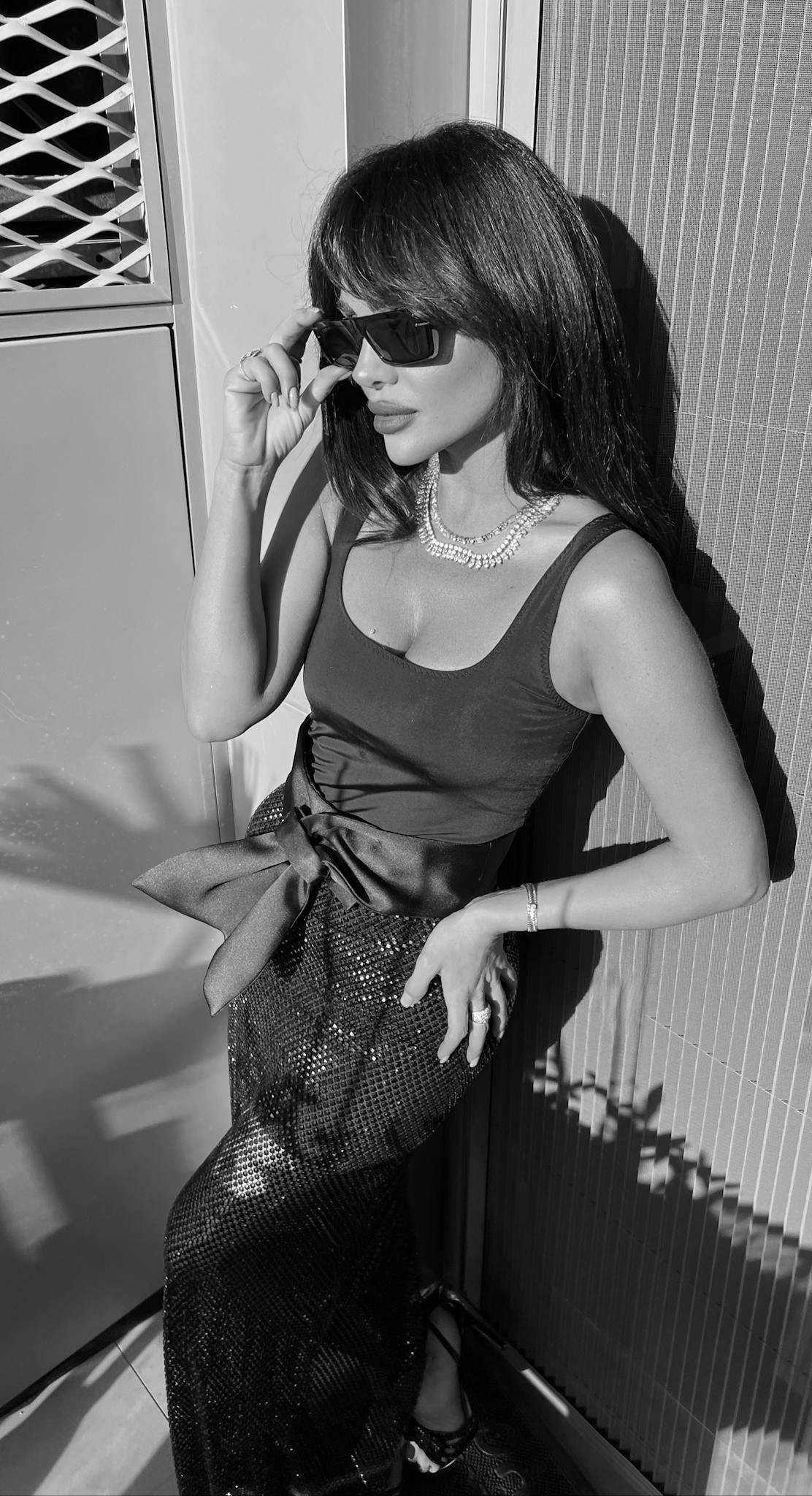 Stylish woman poses in elegant attire wearing sunglasses and a necklace.